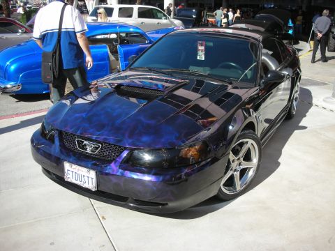 1998 Ford Crown Victoria - black Ford Mustang with swirling purple-and-blue flame paint, custom wheels and an ETDUSTT plate displayed at a sunny car show.