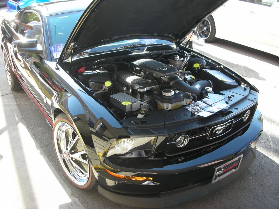 Black Ford Mustang engine bay with supercharger and chrome wheels, hood open