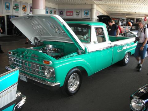 1998 Ford Crown Victoria - green-and-white classic Ford pickup with chrome wheels and open hood revealing the engine, displayed indoors at a car show.