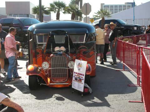 1998 Ford Crown Victoria - orange flame-painted hot rod hauler with twin spotlights and a chrome grille parked behind red barriers at an outdoor show.