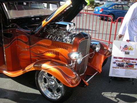 1998 Ford Crown Victoria - orange flame-painted hot rod with a chrome blown engine, polished wheels and open hood, displayed beside an info placard outdoors.