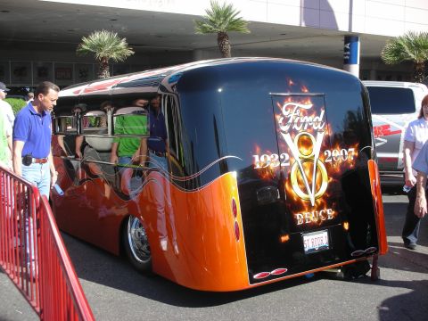 1998 Ford Crown Victoria - Streamlined black and orange custom bus with a flaming Ford V8 Deuce 1932-2007 logo on the rear, parked at an outdoor event with onlookers.