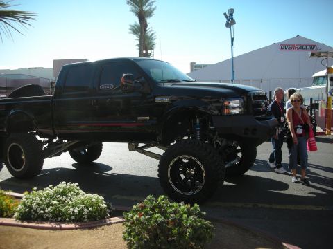 1998 Ford Crown Victoria - Black lifted Ford Super Duty pickup raised on large off-road tires, displayed near an Overhaulin tent with people standing alongside.