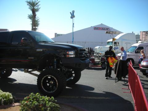1998 Ford Crown Victoria - Front of a black lifted pickup truck on oversized tires and black wheels, parked outdoors near an Overhaulin tent and trailers.