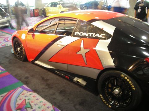 1998 Ford Crown Victoria - Side view of an orange, silver and black race car marked Pontiac with star graphics on Hoosier slicks, displayed indoors.