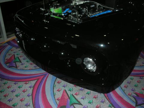 1998 Ford Crown Victoria - Front view of a glossy black custom car with a single round headlight and an exposed green-accented engine, displayed indoors on patterned…