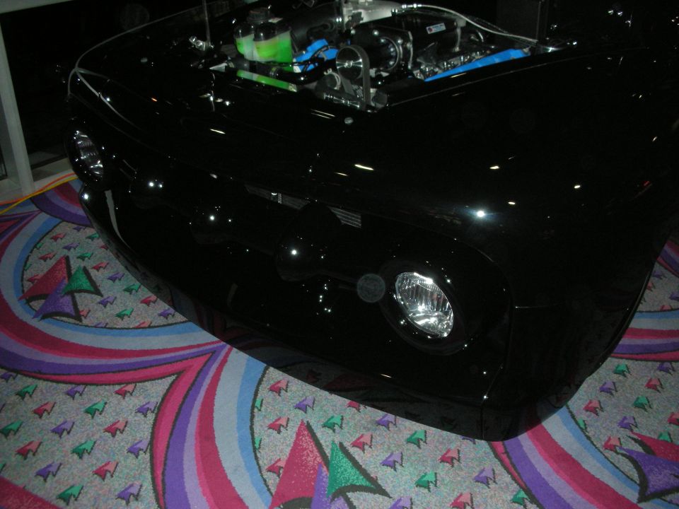 Front of a glossy black custom car with single round headlight and open engine, indoors