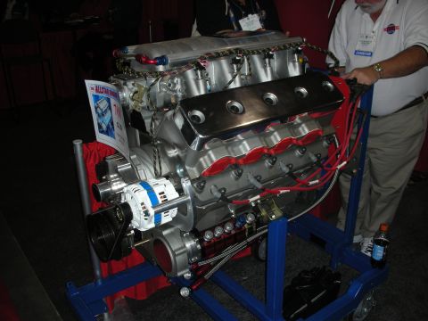 1998 Ford Crown Victoria - large polished aluminum V8 engine mounted on a blue stand at an indoor expo, with red plug wires and a chrome alternator.