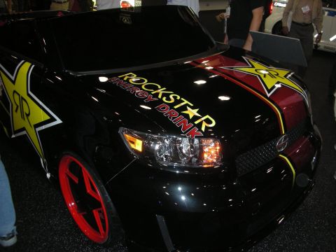 1998 Ford Crown Victoria - black compact car wrapped with yellow Rockstar Energy Drink star graphics and red five-spoke wheels at an indoor show.