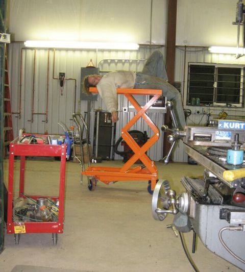 1990 Ford Bronco - A person takes a break on a hydraulic lift in a workshop environment filled with tools and equipment.