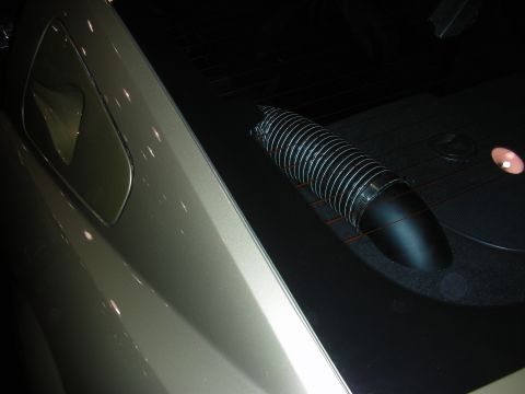 1998 Ford Crown Victoria - ribbed flexible air duct curving into a dark carpeted vehicle cargo area next to a body panel under dim lighting.