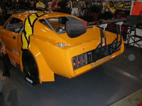 1998 Ford Crown Victoria - yellow-orange wide-body race coupe with a tall carbon-fiber rear wing and Toyo tires displayed at an indoor automotive show.