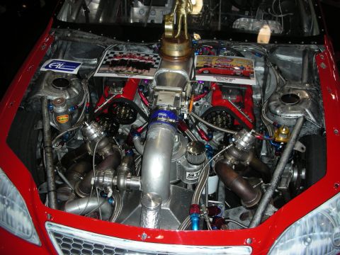 1998 Ford Crown Victoria - engine bay of a red front-drive car packed with a twin-turbo build, braided fuel lines and a forest of plumbing.