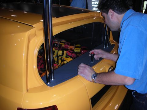 1998 Ford Crown Victoria - man in a blue polo leans into the trunk of a yellow custom car, examining a control near a tray of patterned items inside.