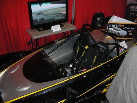 1998 Ford Crown Victoria - black and silver open-cockpit track car with twin Recaro bucket seats parked at a booth next to a racing-video screen and a sweepstakes box.