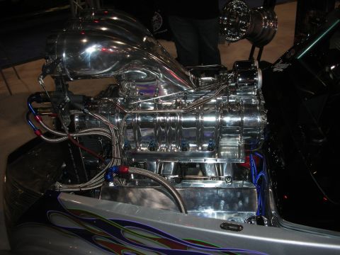 1998 Ford Crown Victoria - polished chrome supercharged V8 engine sits in a custom car with flame-painted bodywork at an indoor show.