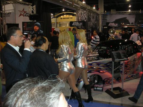 1998 Ford Crown Victoria - Crowded indoor automotive trade-show aisle with two promotional models in silver outfits beside a flame-painted custom car.