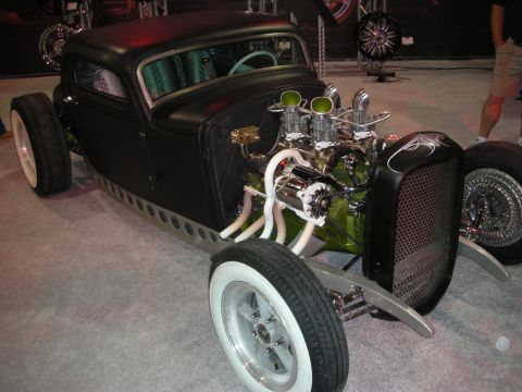 1998 Ford Crown Victoria - Chopped flat-black rat rod on whitewall tires with an exposed dual-carburetor V8 and white headers at a show.