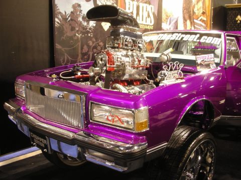 1998 Ford Crown Victoria - Purple custom car with its hood up revealing a chrome supercharged V8 engine, displayed against a graphic backdrop.