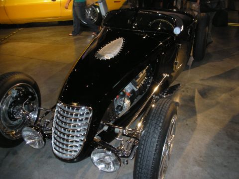 1998 Ford Crown Victoria - Black vintage-style boattail roadster with a chrome grille, exposed suspension and chrome wire wheel on a concrete floor.