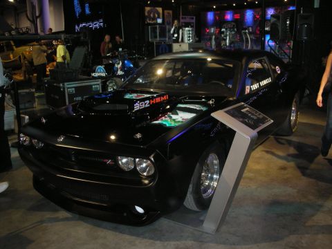 1998 Ford Crown Victoria - black Dodge Challenger marked Test Vehicle sits on a show stand, its 392 HEMI hood scoop and chrome drag wheels lit by booth lighting.