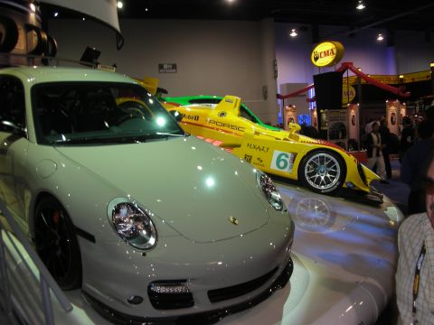 1998 Ford Crown Victoria - white Porsche 911 sits in the foreground while a yellow Mobil 1 Porsche prototype racer is displayed behind it under booth lights.