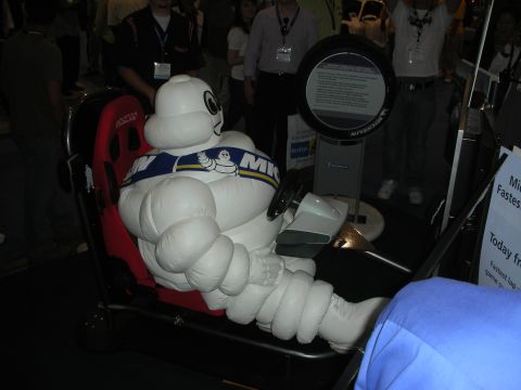 1998 Ford Crown Victoria - inflatable Michelin Man mascot sits behind the wheel of a driving simulator rig as attendees watch a tire-themed display.
