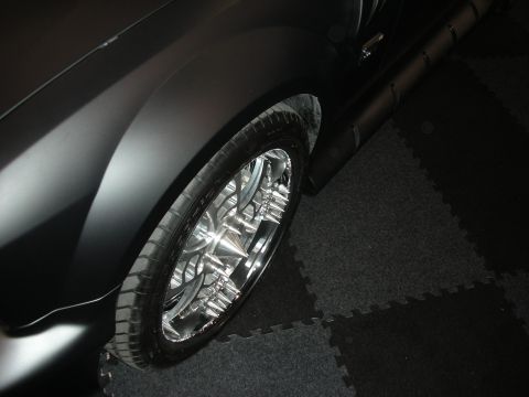 1998 Ford Crown Victoria - polished chrome multi-spoke wheel sits under the dark fender of a matte-black car parked on interlocking display mats.