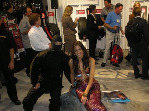 1998 Ford Crown Victoria - person in a black ninja outfit crouches beside a model in a sequined mermaid costume on a fake rock at a busy tire trade-show booth.