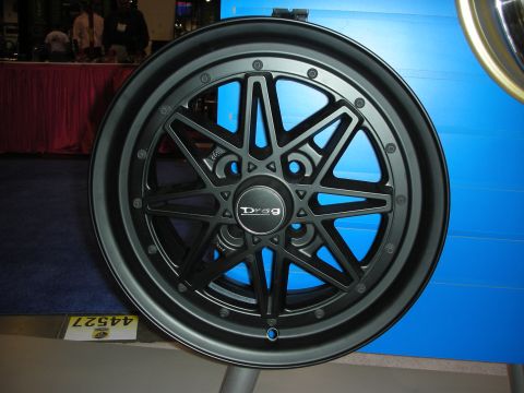 1998 Ford Crown Victoria - matte-black Drag-brand multi-spoke alloy wheel sits on a display stand against a blue booth wall.