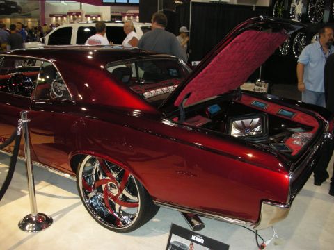 1998 Ford Crown Victoria - deep-red custom muscle coupe sits behind velvet ropes with its trunk open to reveal a tufted audio install and big chrome wheels.