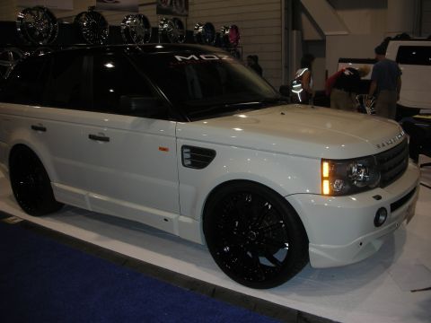 1998 Ford Crown Victoria - white Range Rover Sport with glossy black wheels sits beside a wall of display wheels at an indoor show.