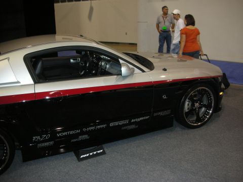 1998 Ford Crown Victoria - black-and-silver Ford Mustang with a red pinstripe and chrome multi-spoke wheels sits at an auto show, its lower body covered in sponsor de…