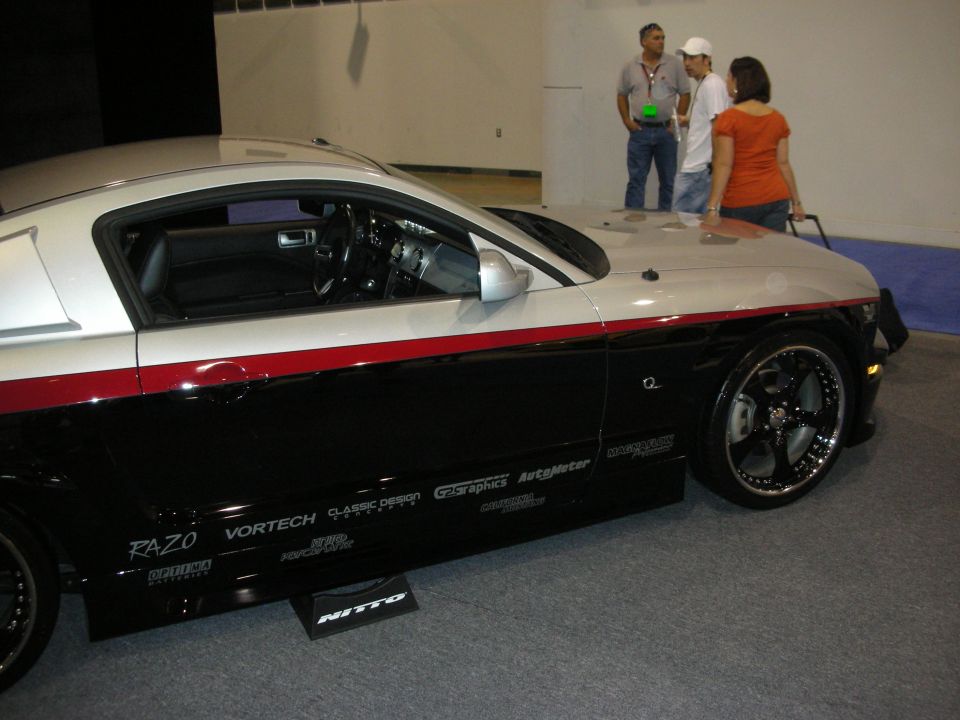 Black and silver Mustang coupe with red pinstripe and chrome wheels at an auto show, sponsor decals on lower door