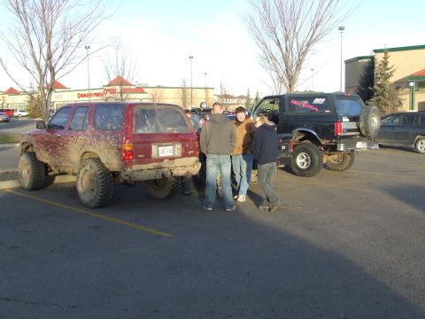 1990 Ford Bronco - Two off-road vehicles are parked in a lot, one covered in mud, with a group of people gathered nearby.