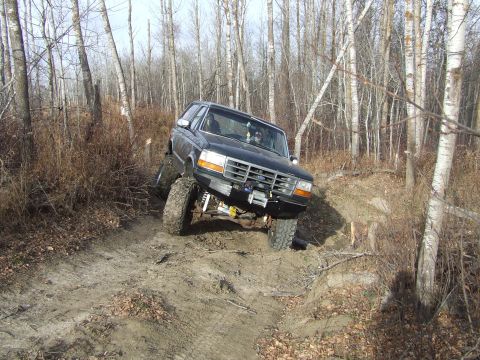 1990 Ford Bronco - A black Ford Bronco navigates a challenging off-road trail in a wooded area.