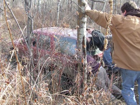 1990 Ford Bronco - A red vehicle is seen partially hidden by trees and brush, with several people gathered nearby.