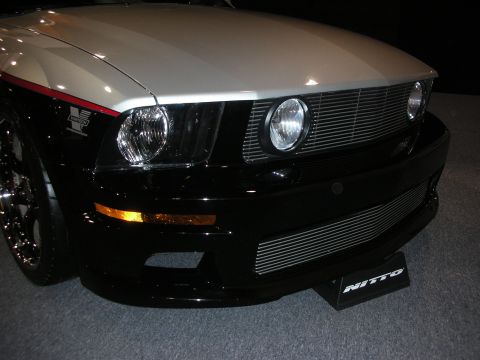1998 Ford Crown Victoria - Front corner of a black-and-silver Ford Mustang showing a billet mesh grille, round headlights and chrome wheels at an auto show.