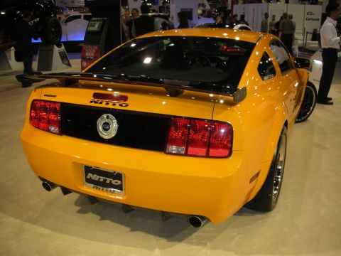 1998 Ford Crown Victoria - Rear three-quarter view of an orange Ford Mustang coupe with a spoiler and dual exhaust on a show stand marked Nitto.