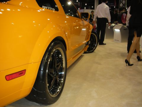 1998 Ford Crown Victoria - Low side view of an orange Mustang showing its black multi-spoke wheels and fender at a busy auto show.