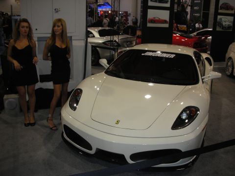 1998 Ford Crown Victoria - white Ferrari coupe displayed with two women in black dresses beside it at a crowded auto show.