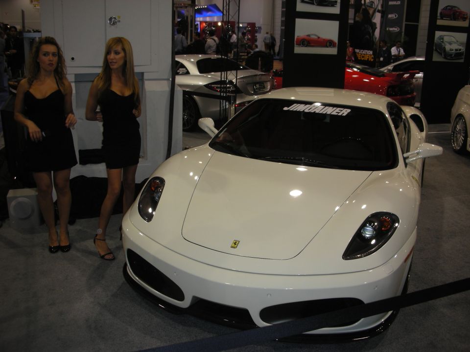 White Ferrari coupe with two women in black dresses standing beside it at an auto show