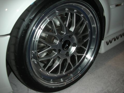 1998 Ford Crown Victoria - Close-up of a silver multi-spoke wheel with a polished lip and bolts on a white car at an auto show.