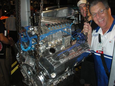 1998 Ford Crown Victoria - Two smiling men pose beside a polished chrome Mopar Hemi V8 crate engine suspended on chains at an indoor event.
