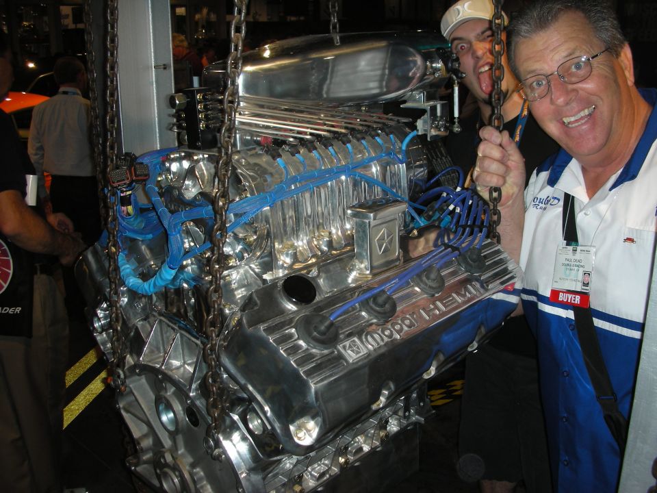 Two men posing with a polished chrome Mopar Hemi V8 engine hanging on chains at a show