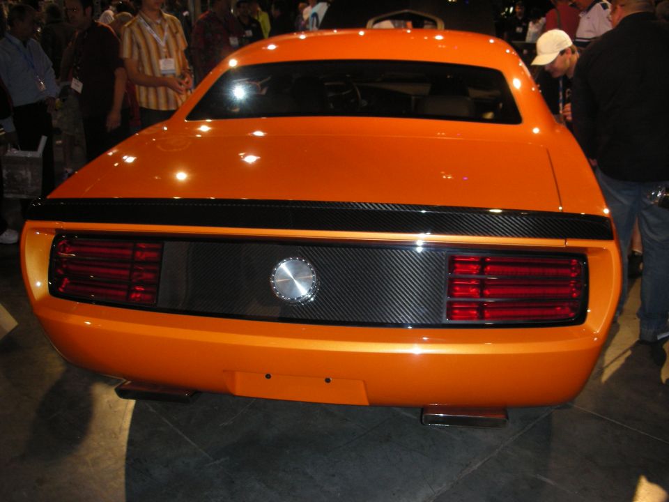 Rear of an orange muscle-style coupe with carbon panel and round taillights in a show crowd