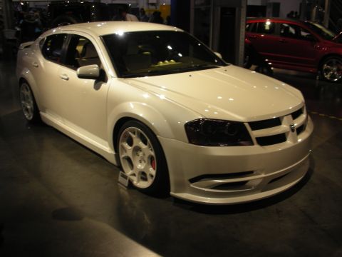 1998 Ford Crown Victoria - customized white Dodge sedan with a body kit and white wheels sits on a dark auto show floor near a red car.