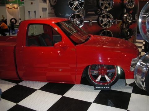 1998 Ford Crown Victoria - bodydropped red pickup truck on chrome wheels and Nitto tires displayed on a checkered floor in front of a wheel wall.