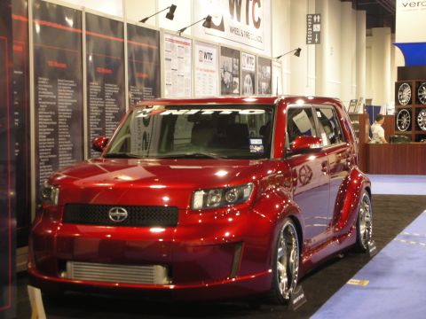 1998 Ford Crown Victoria - candy-red Scion xB sits lowered on chrome wheels at an indoor WTC wheel-and-tire trade-show booth.