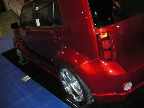 1998 Ford Crown Victoria - Rear three-quarter view of a candy-red Scion xB lowered on chrome wheels, parked on a black mat at an indoor show.